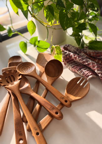 A set of wooden kitchen utensils, including spoons, a fork, and a slotted spoon, arranged on a light surface with a green plant and a patterned cloth in the background.