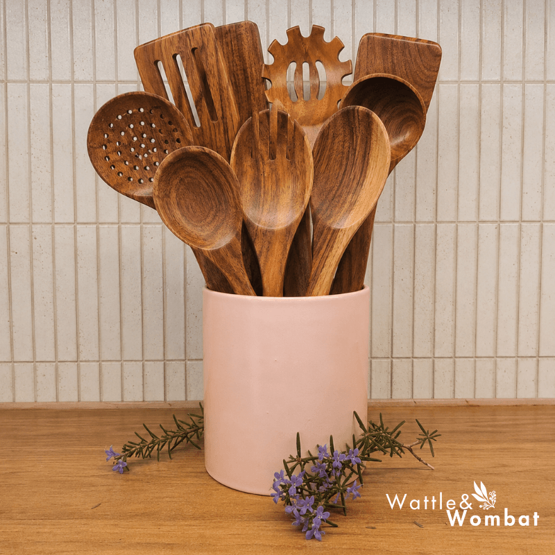 A set of wooden kitchen utensils, including spoons, spatulas, and a pasta server, arranged in a light pink ceramic holder on a wooden surface with small purple flowers in front. The background shows a beige tiled wall.
