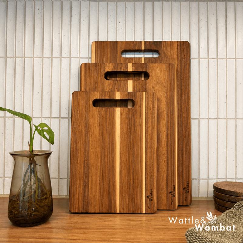 Three wooden cutting boards of different sizes with integrated handles, arranged in a stack on a wooden countertop against a white subway tile wall. A glass vase with a green plant is on the left, and a stack of round wooden items is on the right.