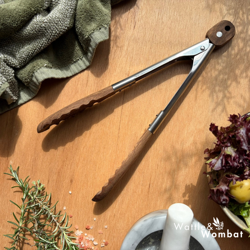 Pair of tongs on a wooden surface with herbs and a mortar and pestle.