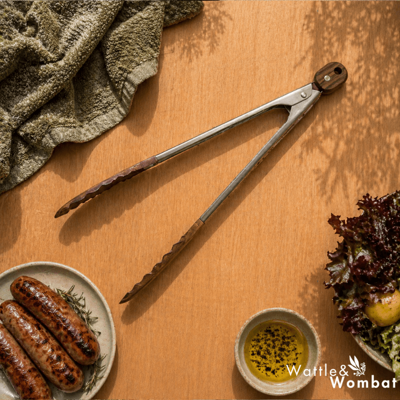 Metal tongs on a wooden surface with sausages, a bowl of oil, and a green towel.