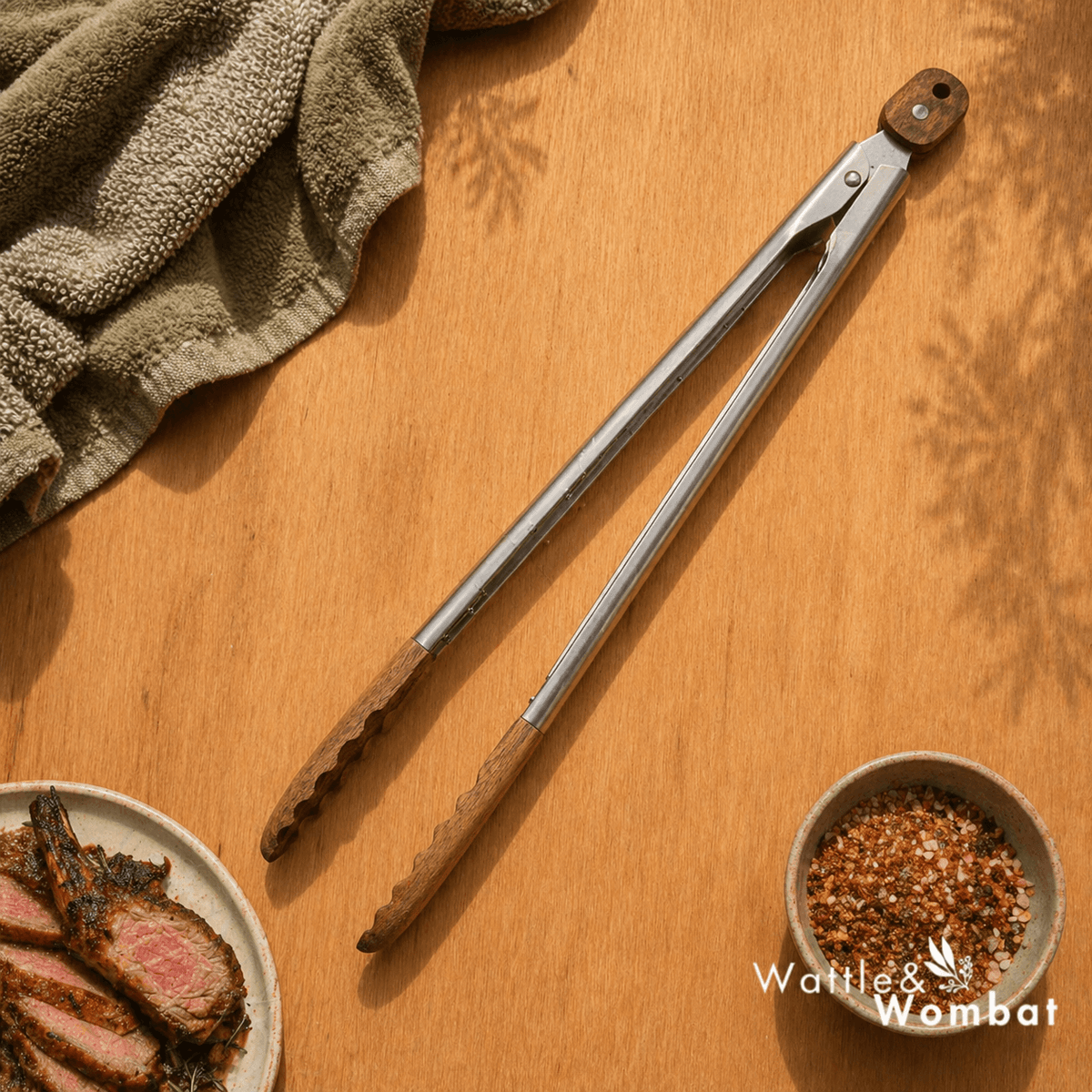 Metal tongs on a wooden surface with a plate of sliced meat and a bowl of spices, featuring the brand 'Wattle & Wombat'.